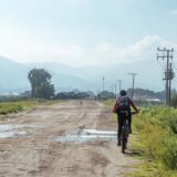 Biciclist cu cască pedalând pe un drum asfaltat de țară, printre câmpuri verzi