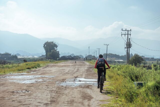 Biciclist cu cască pedalând pe un drum asfaltat de țară, printre câmpuri verzi