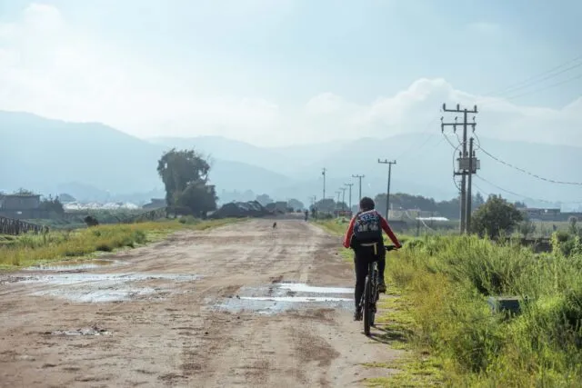 Biciclist cu cască pedalând pe un drum asfaltat de țară, printre câmpuri verzi