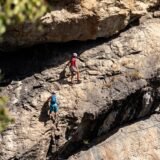 Turist cu cască și ham care urcă pe un perete de stâncă asigurat de cablul de oțel al unei via ferrata
