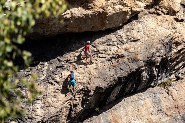 Turist cu cască și ham care urcă pe un perete de stâncă asigurat de cablul de oțel al unei via ferrata